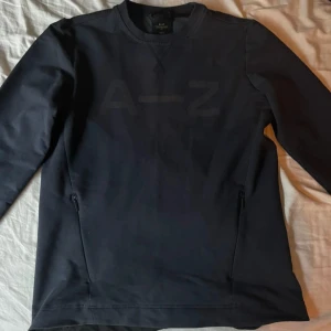 Svart sweatshirt från A-Z med dragkedjor - Säljer en svart sweatshirt från A-Z i storlek M. Tröjan har diskret A-Z-logga framtill, rund halsringning och två praktiska dragkedjor vid sidorna. Perfekt för en clean och stilren look.