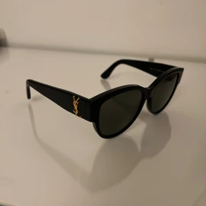 Svarta ysl solglasögon - Snygga svarta solglasögon från Saint Laurent med klassisk cat-eye form och mörka linser. Tjocka bågar i blank plast med ikonisk YSL-logo i guld på sidorna. Perfekt accessoar för att höja din stil och ge en lyxig vibe.