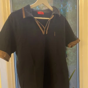 Svart Hugo Boss henley med bruna detaljer - Svart henley-skjorta från Hugo Boss med bruna kontrastdetaljer på krage och ärmslut. Kortärmad modell med v-ringad hals och knappar. Diskret Hugo-logga på bröstet. Perfekt för dig som gillar stilrena plagg med en twist.