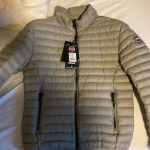 Beige pufferjacka från Colmar - Snygg beige pufferjacka från Colmar med horisontella sömmar och hög krage. Jackan har två dragkedjeförsedda fickor och en diskret logotyp på ärmen. Perfekt för kyliga dagar och enkel att matcha med olika outfits.