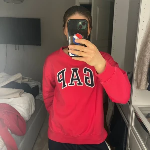 Röd GAP sweatshirt med logga - Röd sweatshirt från GAP med stor vit och svart logga framtill. Klassisk rund halsringning och ribbade muddar vid ärmslut och nederkant. Skön och avslappnad passform, perfekt till jeans eller mjukisbyxor. Materialet är mjuk bomullsmix.