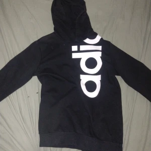 Svart Adidas hoodie med stor logga - Svart hoodie från Adidas med stor vit logga som går diagonalt över både fram- och baksidan. Klassisk huva och långa ärmar med ribbade muddar. Perfekt för dig som gillar sportig och street-inspirerad stil.