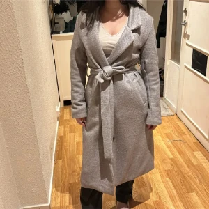 Grå lång kappa med bälte - Säljer en stilren grå kappa i lång modell med bälte i midjan. Kappan har klassisk krage, raka ärmar och två fickor framtill. Perfekt för dig som vill ha en enkel men snygg look under höst och vinter. Tror tror den är från lager157 men köpt för flera år sen men fint skick kan finnas lite noppror men inget störande💕