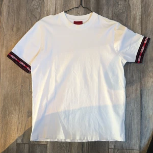 Vit t-shirt från Hugo Boss med logga - Vit t-shirt från Hugo Boss med klassisk passform. Ärmarna har svarta och röda detaljer med Hugo-logga. Tillverkad i mjuk bomull och har rund halsringning. Perfekt för en clean och stilren look.
