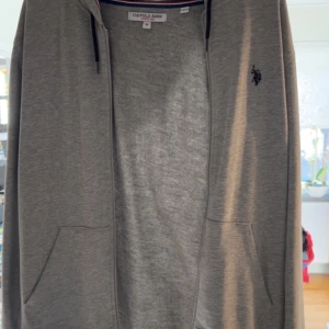 Grå hoodie från U.S. Polo Assn M - Snygg grå hoodie från U.S. Polo Assn med dragkedja, huva med svarta snören och två fickor framtill. Klassisk logga broderad på bröstet och mjukt bomullsmaterial. Perfekt för chill dagar eller när du vill ha en enkel streetstyle.