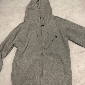 Grå hoodie från Ralph Lauren - Säljer en stilren grå hoodie från Ralph Lauren med dragkedja och huva. Tröjan har snörning i huvan, lång ärm och den klassiska broderade loggan på bröstet. Perfekt för en chill och avslappnad look.