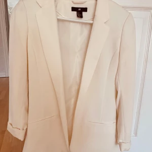 Vit kavaj från H&M, storlek 36 - Snygg vit kavaj från H&M i storlek 36. Klassisk modell med smal krage, två fickor fram och helfodrad insida. Stilren och enkel design som ger en fräsch look. Perfekt att matcha med jeans eller kostymbyxor för en clean vibe.
