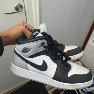Nike Air Jordan 1 Mid svart/vit/grå - Säljer ett par Nike Air Jordan 1 Mid i svart, vitt och grått skinn. Klassisk siluett med svart swoosh, svarta snören och ikoniska Air Jordan-loggan på sidan. Perfekta för dig som gillar streetstyle och vill ha en sneaker med hype-känsla. Priset kan diskuteras 