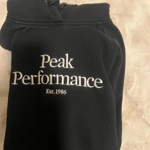 Svart hoodie från Peak Performance - Svart hoodie från Peak Performance med vit broderad logga på bröstet och klassisk känguruficka. Tröjan har huva med snörning och är perfekt för en chill och sportig stil. Mjuk insida och relaxed fit.