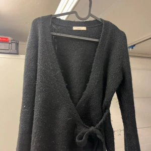 Svart omlottkofta från Gina Tricot - Mysig svart omlottkofta från Gina Tricot med knyt i sidan. Koftan är stickad och har lång ärm samt djup v-ringning. Perfekt för lager-på-lager och passar till allt. Enkel och stilren design som funkar året om.