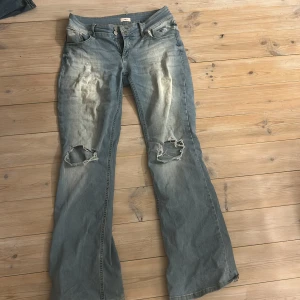 Ljusblå bootcut jeans från Nelly - Säljer ett par ljusblå jeans från Nelly med bootcut-modell. Jeansen har slitna detaljer och stora hål vid knäna för en cool, avslappnad look. Klassiska femficksdetaljer och dubbla knappar framtill. Perfekta för dig som gillar en edgy stil.