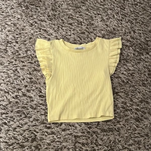 Gul ribbad topp med volangärm från Zara - Supersöt ljusgul ribbad topp från Zara med volangdetaljer på ärmarna. Toppen är ärmlös med rund halsringning och croppad passform. Ganska kort i längden. Perfekt för vår och sommar när du vill ha något färgglatt och bekvämt. 