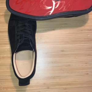 Christian Louboutin svarta sneakers mocka - Svarta sneakers från Christian Louboutin i lyxig mocka med klassisk röd sula och diskret logga. Skorna har rund tå, snörning och en stilren design som sticker ut med den ikoniska röda undersidan.