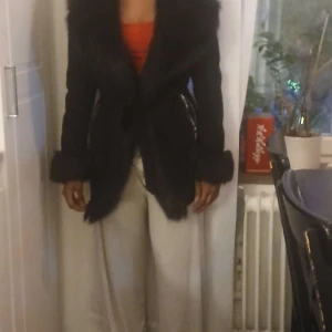 Svart pälsjacka med bälte - Säljer en svart pälsjacka med bred krage och bälte i midjan. Jackan har långa ärmar och dragkedjor vid fickorna som ger en snygg detalj. Perfekt för dig som vill ha en varm och stilren look under kyliga dagar.