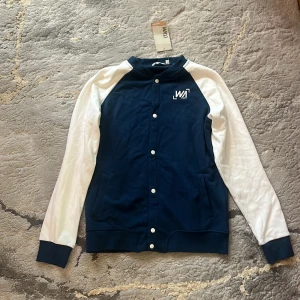Blåvit collegejacka från WA - Snygg blåvit collegejacka från WA med vita ärmar och marinblå kropp. Tryck med WA-logga på bröstet och ryggen, knäppning med vita knappar och två fickor framtill. Perfekt för en sportig och avslappnad stil.