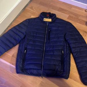 Mörkblå pufferjacka med gul detalj - Säljer en mörkblå parajumper jacka med horisontella sömmar, hög krage och en snygg gul knappdetalj vid halsen. Jackan har dragkedja framtill, tre fickor med dragkedja och en modern, sportig look. Perfekt för kyliga dagar.