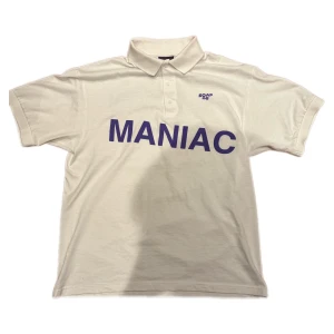 Vit polo från Soap08 med tryck - Vit piké från Soap08 med lila tryck 'MANIAC' på framsidan och 'Malibu Beach Nightmare BE MY VICTIM' på ryggen. Klassisk krage och knappar framtill. Tillverkad i 100% bomull, made in Los Angeles. Statement-plagg med edgy vibe.