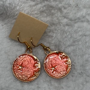 Guldiga örhängen med månansikte - Unika örhängen i guldton med rund medaljongform, dekorerade med ett ansikte i profil och en stjärna. Färgen är en mix av guld och korallrosa, vilket ger en cool och trendig vibe. Perfekta för dig som vill sticka ut med accessoarer som har en konstnärlig touch.