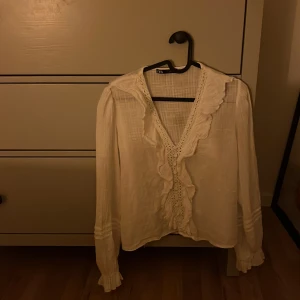 Vit blus med volang från Zara - Supersöt vit blus från Zara med v-ringning, volangdetaljer och spets framtill. Långärmad med lätt puff vid axlarna och rynkade muddar. Luftigt material som känns skönt mot huden och ger en romantisk vibe. Perfekt för dig som gillar detaljer och vill sticka ut lite extra. Jag har aldrig använt den men köpt från vinted sen tidigare så kan vart använd hos den förra ägaren🥰