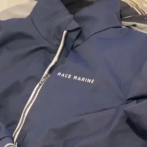 Blå vindjacka från Race Marine - Snygg marinblå vindjacka från Race Marine med vita dragkedjor och hög krage. Jackan har raka linjer, praktiska fickor och är perfekt för blåsiga dagar. Enkel och stilren design som funkar till det mesta.