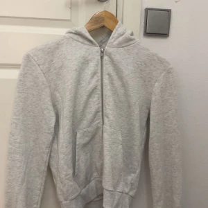 Ljusgrå hoodie med dragkedja - Enkel och stilren ljusgrå hoodie med dragkedja framtill och huva. Tröjan har långa ärmar och praktiska fickor på magen. Perfekt för en avslappnad look och passar till det mesta.