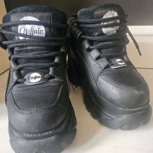 Buffalo London svarta platåsneakers - Helt nya Buffalo London svarta sneakers med chunky platåsula och rund tå. Skorna har snörning framtill, tydlig logga på plös och häl samt ovandel i skinn och foder i polyester. Perfekta för dig som gillar 2000-talsvibbar och vill sticka ut med en rejäl sula.