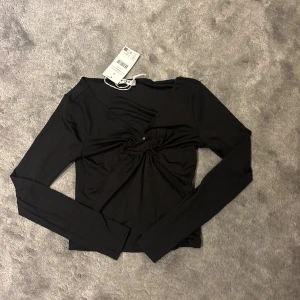 Svart långärmad topp från NA-KD - Svart långärmad topp från NA-KD med v-ringning och snygg rynkdetalj framtill som hålls ihop av en guldfärgad metallring. Toppen har en figurnära passform och är tillverkad i mjukt, stretchigt material. Perfekt för att lyfta din outfit med en cool detalj.