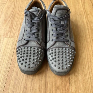 Grå Christian Louboutin sneakers med nitar - Säljer ett par gråa sneakers från Christian Louboutin i mocka med ikoniska röda sulor. Skorna har snörning och coola nitar på tån för en edgy vibe. Insidan är i lyxigt ljust skinn och detaljerna är riktigt snygga. Perfekta för dig som vill sticka ut.