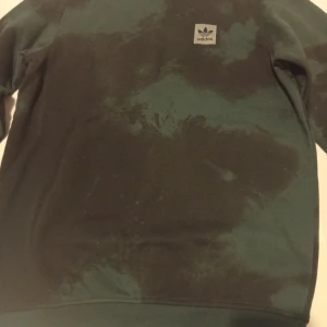 Mörkgrön crewneck från Adidas - Säljer en mörkgrön crewneck från Adidas med batikmönster i svart. Tröjan har rund hals, långa ärmar och en vit Adidas-logga på bröstet. Perfekt för dig som gillar streetwear och vill ha en unik look.