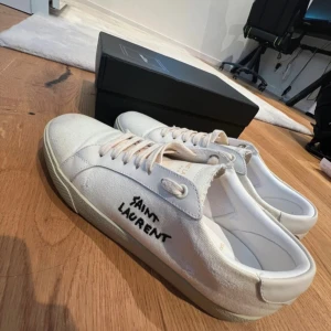 Saint Laurent skor - Snygga vita sneakers från Saint Laurent med handskriven logga på sidan. Skorna har rund tå, snörning och är gjorda i canvas med detaljer i skinn. Klassisk platt sula och stilren design som passar till allt. Kommer med originalkartong och dustbag.