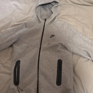 Nike tech grå storlek s - Säljer en stilren ljusgrå Nike hoodie i storlek S. Tröjan har hel dragkedja, svarta detaljer vid fickorna och klassisk Nike-logga på bröstet. Mjuk och skön med huva, perfekt för chill eller träning.
