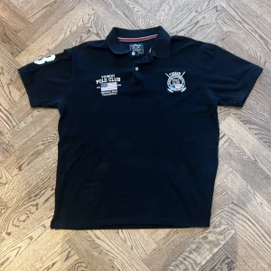 Svart pikétröja Vinson Polo Club - Svart pikétröja från Vinson Polo Club med klassisk krage och knappar framtill. Broderade loggor på bröstet och tryckt nummer på ärmen. Skön passform och stilren look, perfekt till jeans eller shorts. 