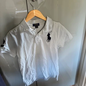 ljusblå pikétröja från Polo Ralph Lauren - Ljusblå pikétröja från Polo Ralph Lauren med klassisk krage, korta ärmar och stor marinblå logga på bröstet. Snygg detalj med en stor siffra 3 på ena ärmen. nypris 1000 kr