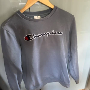 Blågrå sweatshirt från Champion XXS - Säljer en blågrå sweatshirt från Champion med stort broderat logotyptryck över bröstet. Tröjan har rund halsringning, långa ärmar och är tillverkad i mjuk bomullsmix. nypris 550 kr
