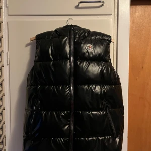 Svart dunväst från Moncler - Snygg svart dunväst från Moncler med glansig finish och klassisk logga på bröstet. Västen har huva, två dragkedjefickor och hel dragkedja framtill. Perfekt för lager-på-lager och streetstyle.