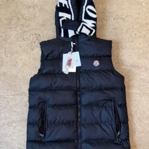  Moncler väst  - Snygg svart dunväst från Moncler med avtagbar huva som har svartvita detaljer och logotyp. Västen har två dragkedjefickor framtill och klassisk Moncler-logga på bröstet. Perfekt för lager-på-lager och streetstyle.