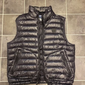 Svart Glossed Down Vest från Polo Ralph Lauren - Snygg svart dunväst från Polo Ralph Lauren med quiltad design och hög krage. Västen har två dragkedjeförsedda fickor framtill och diskret logga på bröstet. Perfekt för lager-på-lager och enkel att matcha med olika outfits. Som ny