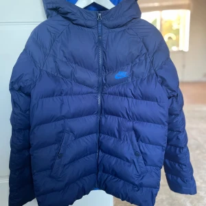 Blå vinterjacka från Nike - Blå vinterjacka junior, 13-15 år, från Nike med huva och dragkedja framtill. Jackan har två fickor med knappar och är vadderad. Endast använd 2 ggr. 