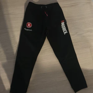 Svarta mjukisbyxor från Hoodrich  - Svarta mjukisbyxor från Hoodrich med tryckt logga på benet och röd detalj på snörningen. Byxorna har elastisk midja med dragsko och är tillverkade i mjukt material för maximal komfort. Snygga detaljer med broderad signatur och röd/vit logotyp på låret.