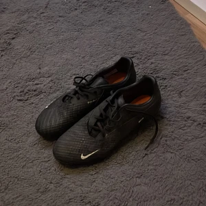 Nike Phantom GT2 svarta fotbollsskor - Säljer ett par svarta Nike Phantom GT2 fotbollsskor med orange dobbar och vit swoosh på sidan. Skorna har snörning och är tillverkade i syntetmaterial med en rund tå och platt sula. Perfekta för dig som vill ha grym kontroll på planen.