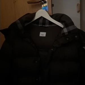 Svart pufferjacka Burberry XL - Svart pufferjacka från Burberry i storlek XL. Jackan har klassisk Burberry-rutig insida på kragen, dragkedja framtill och en snygg patch med Burberry-logga på ärmen. Perfekt för kalla dagar med sin tjocka och fluffiga design.