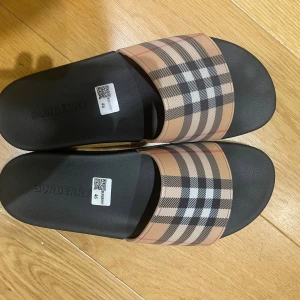 Burberry tofflor beige med rutig rem - Säljer ett par svarta sandaler från Burberry med den klassiska beige och svarta rutiga remmen över foten. Snyggt präglad Burberry-logga på fotbädden och undersidan. Tillverkade i Italien, gjorda i syntetmaterial med platt sula och öppen tå.
