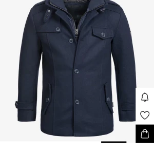 Mörkblå rock från INDICODE - Snygg mörkblå rock från INDICODE med klassisk krage, knappar framtill och dragkedja. Jackan har fickor med lock, axelklaffar och detaljer vid ärmslut. Perfekt för dig som vill ha en stilren och tidlös look under höst och vinter. (Knappen längs ner har gått sönder och är borta)