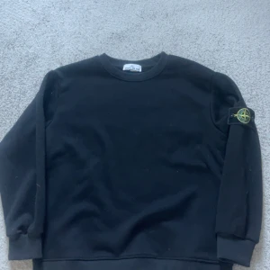 Svart sweatshirt från Stone Island - Svart sweatshirt från Stone Island med klassisk rund hals och ribbade muddar. Ikonisk logotyp-patch på vänster ärm. Tröjan är i mjuk bomull och har en clean, stilren look som passar perfekt till streetwear.tröjan är inte äkta