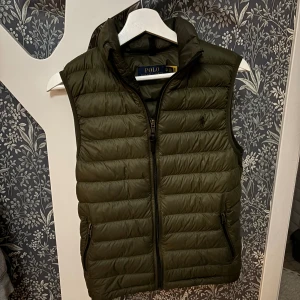 Olivgrön dunväst från Polo Ralph Lauren - Snygg olivgrön dunväst från Polo Ralph Lauren med quiltad design och dragkedja framtill. Västen har hög krage och två fickor med dragkedja. Perfekt att slänga över en hoodie eller tröja för extra värme.