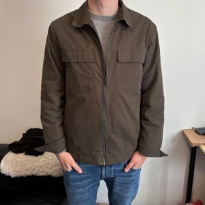 Olivgrön overshirt - Säljer denna stilrena olivgröna overshirt från Dressmann med dragkedja och två bröstfickor. Jackan är fodrad och varm vilket gör att den funkar bra nu mot vintern med. Modellen är 181cm lång och väger 72kg. Tveka inte vid frågor och prisförslag.✍️🪖