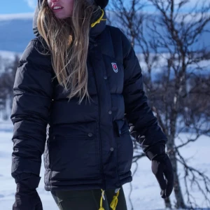 Söker!!! En svart fjällräven jacka till diskussions pris - Svart dunjacka från Fjällräven med huva och gula detaljer vid snörningen. Jackan har en klassisk pufferlook, knappar framtill och två stora fickor. Perfekt för kalla dagar och outdoor-äventyr.