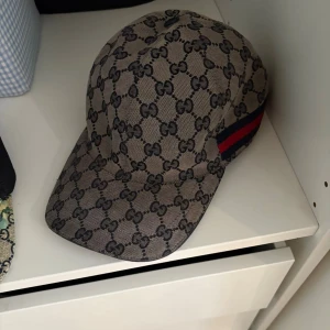 Gucci keps med GG-mönster - Säljer en snygg keps från Gucci med klassiskt GG-monogram i svart på beige bakgrund. Kepsen har en böjd skärm och ikoniska röd-blå ränder på sidan. Tillverkad i canvasliknande material, perfekt för dig som vill sticka ut med en lyxig accessoar.