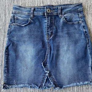 Blå jeanskjol från Pink & Sky - Snygg blå jeanskjol från Pink & Sky med fransad nederkant och klassiska fickor både fram och bak. Kjolen har en kort passform, slitdetaljer och knappstängning framtill. Perfekt för dig som gillar en avslappnad och trendig stil. Mått Midjan: ca 36 cm Total längd: ca 41 cm  68% bomull, 31% polyester och 1% elastan  Kommer från ett djur och rökfritt hem.