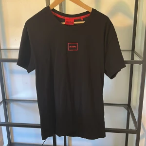 Svart t-shirt från Hugo Boss - Svart t-shirt från Hugo Boss med röd logga på bröstet. Klassisk rund halsringning och korta ärmar. Tillverkad i mjuk bomull för en skön känsla. Enkel och stilren design som passar perfekt till jeans eller shorts.